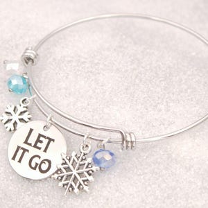 May include: Silver-toned bangle bracelet with snowflake charms and blue crystal accents. A round charm displays the phrase "LET IT GO" in black lettering. The bracelet has a winter theme.