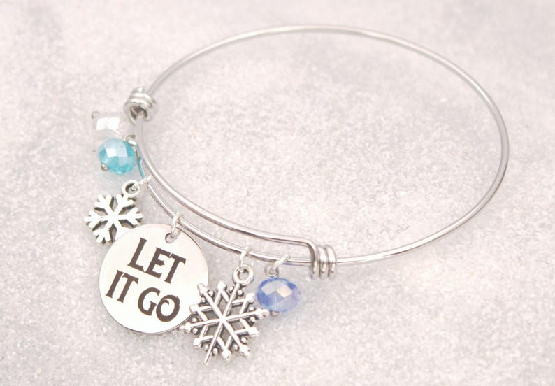 May include: Silver-toned bangle bracelet with snowflake charms and blue crystal accents. A round charm displays the phrase "LET IT GO" in black lettering. The bracelet has a winter theme.
