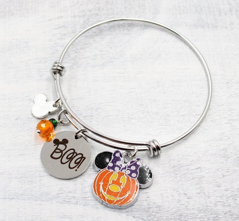 May include: A silver bangle bracelet with charms. The charms include a Mickey Mouse head, orange beads, a round charm with the word "Boo!" and a pumpkin-shaped charm with a Minnie Mouse face.