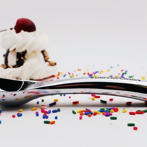Op de afbeelding: Een zilveren ijslepeltje met de woorden "Dad's Ice Cream" gegraveerd op de steel. De lepel ligt op een wit oppervlak met kleurrijke sprinkels eromheen.