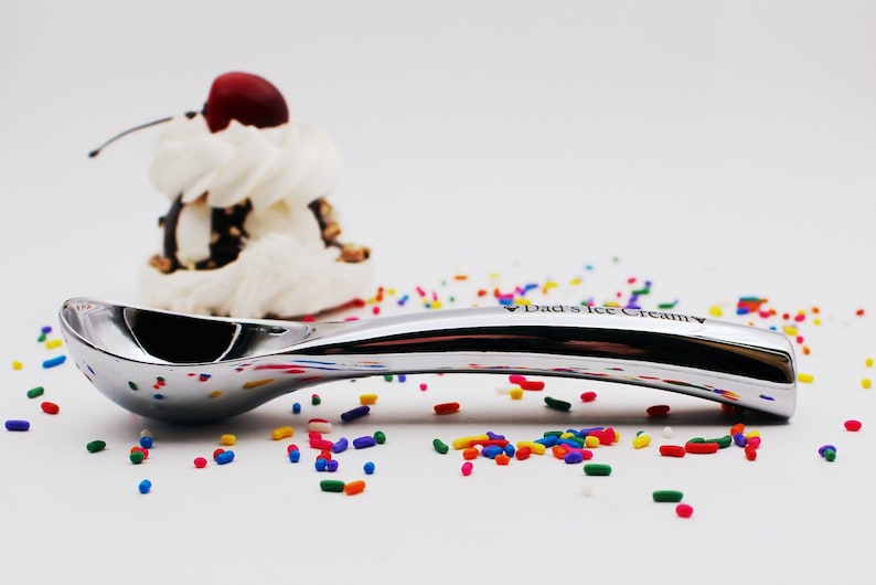 Op de afbeelding: Een zilveren ijslepeltje met de woorden "Dad's Ice Cream" gegraveerd op de steel. De lepel ligt op een wit oppervlak met kleurrijke sprinkels eromheen.