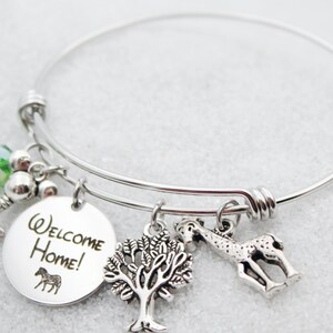 May include: Silver charm bracelet with a round charm that says "Welcome Home!" and features a zebra, a tree charm, a giraffe charm, a key charm, and a green crystal bead.