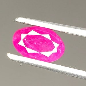 May include: A vibrant, oval-cut ruby gemstone held by silver tweezers. The ruby displays a deep pink hue with a star-like pattern within the stone. The background is a neutral gray, highlighting the gem's color and clarity.