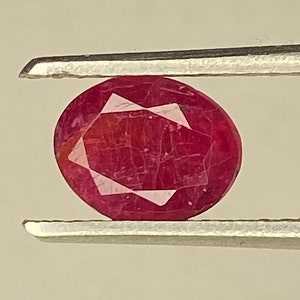 Può includere: Una pietra preziosa ovale di colore rosso scuro tenuta in una pinza. La pietra preziosa ha un taglio sfaccettato ed è traslucida.