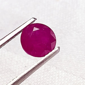 May include: A round, faceted ruby gemstone with a deep pink hue. The ruby is held in a pair of tweezers.