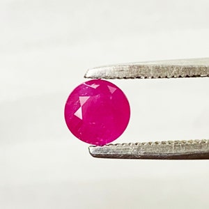 Puede incluir: Una piedra preciosa de rubí redonda y facetada de un tono rosa intenso, sostenida en unas pinzas sobre un fondo blanco.
