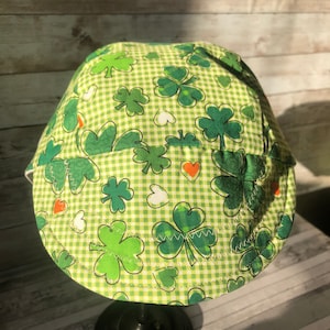 Puede incluir: Un sombrero de tela a cuadros verde y blanco con un patr&oacute;n de tr&eacute;bol. El sombrero est&aacute; en un soporte negro.