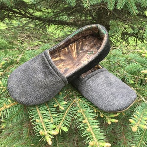 Puede incluir: Un par de zapatillas grises con forro de camuflaje. Las zapatillas están sobre un lecho de agujas de pino verdes.