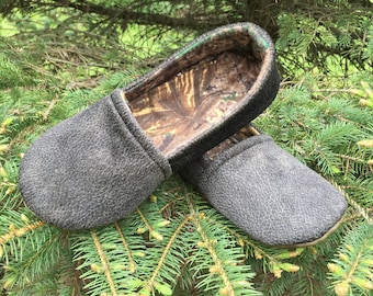 May include: A pair of gray slippers with a camouflage lining. The slippers are sitting on a bed of green pine needles.