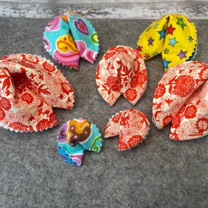 Puede incluir: Una colección de seis juguetes de tela con forma de galleta de la fortuna. Los juguetes están hechos de diferentes telas con varios patrones, incluyendo diseños florales, estrellas y geométricos. Los colores incluyen rojo, blanco, azul, amarillo y verde.