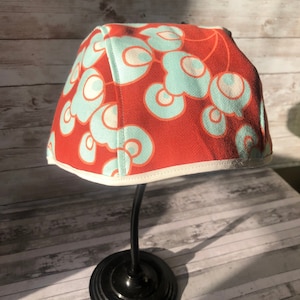 Puede incluir: Un sombrero de tela rojo con ribete blanco y un patr&oacute;n abstracto azul y blanco. El sombrero se muestra en un soporte negro.