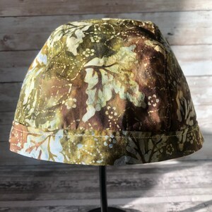 Puede incluir: Un sombrero de tela con estampado verde y marr&oacute;n con ribete azul claro. El sombrero est&aacute; en un soporte negro.