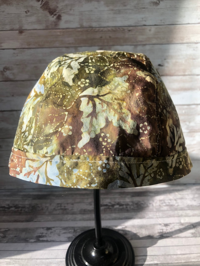 Puede incluir: Un sombrero de tela con estampado verde y marr&oacute;n con ribete azul claro. El sombrero est&aacute; en un soporte negro.