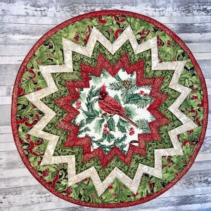 May include: A red cardinal bird with a green holly branch and red berries on a white background. The background is surrounded by a red, green, and white starburst pattern.