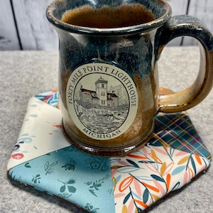 Könnte beinhalten: Eine braune und blaue Keramiktasse mit einem weißen Logo, auf dem "Forty Mile Point Lighthouse Michigan" steht. Die Tasse steht auf einem bunten Stoffuntersetzer.