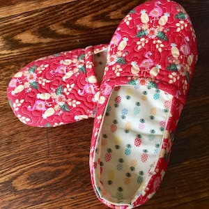 May include: A pair of red quilted slippers with a flamingo and pineapple print. The slippers have a white lining with a pineapple print.