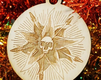 May include: A wooden Christmas ornament with a laser-cut design of a skull inside a flower with radiating lines. The ornament is light brown and has a string for hanging.