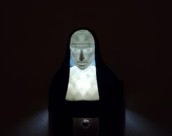 May include: A white night light shaped like a nun with a black habit. The light is plugged into an outlet and is illuminating the face of the nun.