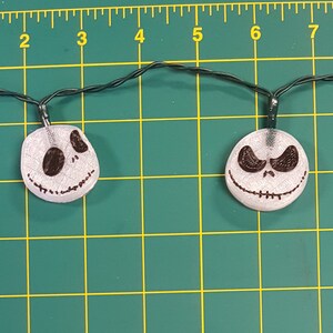 May include: A string of white Christmas lights with Jack Skellington's face on each bulb. The lights are strung on a black wire and are hanging from a green surface.