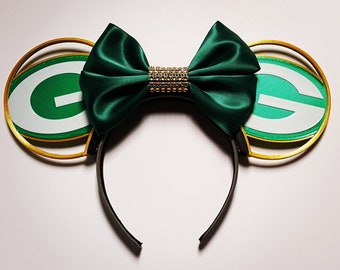 May include: Green and gold Minnie Mouse ears with a green bow and rhinestones. The ears are shaped like the Green Bay Packers logo.