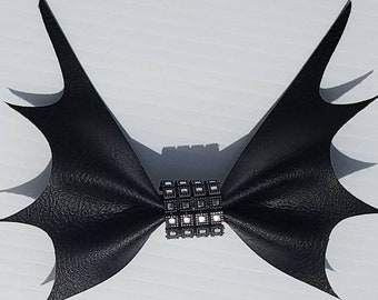 May include: Black leather bow with a spiky, bat-like wing design. The bow is accented with a silver-toned, square-shaped rhinestone embellishment.