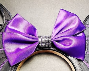 May include: Purple satin bow with a black rhinestone embellishment on a black headband. The headband is designed to look like Mickey Mouse ears.