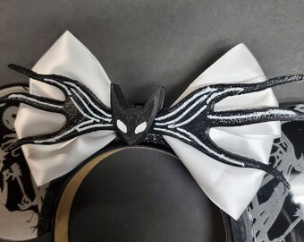 May include: Black and white mouse ears with a 3D printed black bat design with white accents. The bat has a white face with black eyes. The ears are attached to a black headband.