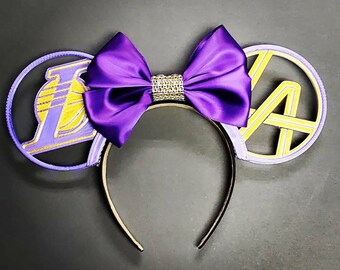 May include: A headband featuring two circular ears in light purple with yellow accents. The left ear displays a purple and yellow basketball logo, while the right ear features a yellow LA monogram. A large, shiny purple satin bow with a rhinestone center sits between the ears.