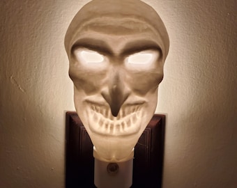 May include: A white ceramic night light shaped like a creepy face with glowing eyes. The night light is plugged into an electrical outlet.