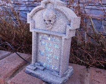 May include: A gray stone tombstone with a skull carved into the top. The tombstone has a message that reads "This is the box we are the cat".