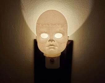 May include: A white porcelain nightlight shaped like a doll's head with glowing eyes. The nightlight is plugged into an electrical outlet.