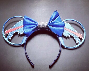 May include: Blue headband with mouse ears featuring a blue satin bow with a rhinestone center. The ears have a blue outline with a Buffalo Bills logo design in red, white, and blue.
