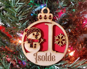 May include: Wooden Christmas ornament with a snowman, snowflake, and the name "Isolde". The ornament has a red background and a natural wood finish. It is hanging on a Christmas tree.