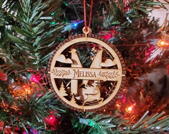 May include: A wooden Christmas ornament with a circular design. The ornament features a mountain scene with a deer, trees, and the name "Melissa" engraved. It hangs from a red string and is displayed on a Christmas tree with colorful lights.