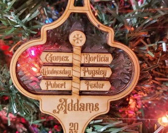 May include: Wooden Christmas ornament with the Addams Family names: Gomez, Morticia, Wednesday, Pugsley, Pubert, and Fester. The ornament has a snowflake and the year 2025. The ornament is hanging on a Christmas tree.