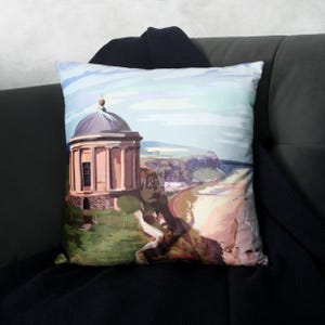 May include: Square cushion with a landscape print depicting a domed building, cliffs, and a beach. The design features a light blue sky and a colour scheme of beige, brown, and green. The cushion is placed on a dark grey sofa.