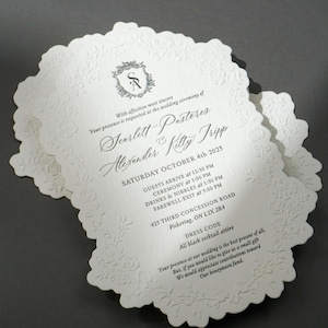 May include: A stack of elegant, scalloped-edge wedding invitations. The ivory cards feature embossed floral designs and include event details, such as the names Scarlett Pastores and Alexander Kitty Fripp, date, time, and location.