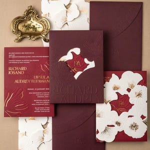 以下が含まれることがあります： エンボス加工されたフローラルデザインとゴールドのアクセントが施された、バーガンディ色のウェディング招待状。招待状セットには、カード、封筒、装飾的な花の切り抜きが含まれています。「Richard」と「Audrey」の名前が大きく表示されています。