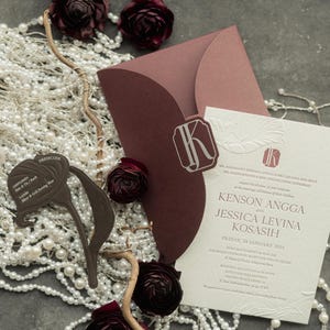 Puede incluir: Un conjunto de invitación de boda con un sobre y una tarjeta de invitación burdeos con detalles en relieve. La invitación incluye los nombres Kenson Angga y Jessica Levina Kosasih. Los adornos incluyen perlas de imitación, flores burdeos y una tarjeta de código de vestimenta.