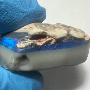 Könnte beinhalten: Ein blauer, grauer und weißer Opal mit einer rauen, natürlichen Textur. Der Opal hat einen blauen, irisierenden Schimmer.