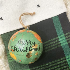 May include: A green globe ornament with a gold map and the words "Merry Christmas" written in black. The ornament is hanging from a stocking with a black and green plaid pattern.