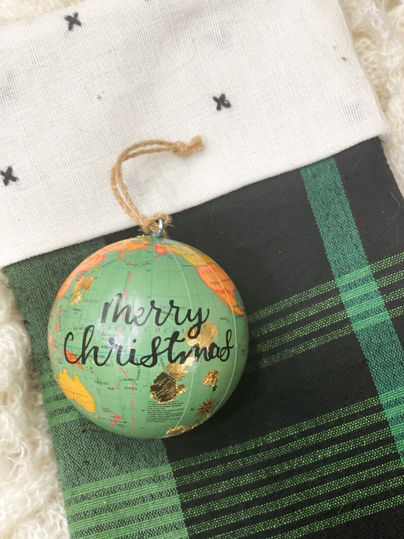 May include: A green globe ornament with a gold map and the words "Merry Christmas" written in black. The ornament is hanging from a stocking with a black and green plaid pattern.