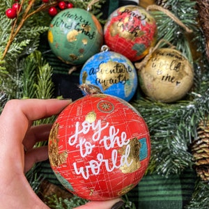 May include: Four red, green, blue, and gold globe ornaments with gold accents and white calligraphy. The ornaments read "Merry Christmas", "Peace on Earth", "Adventure Awaits", and "Joy to the World".