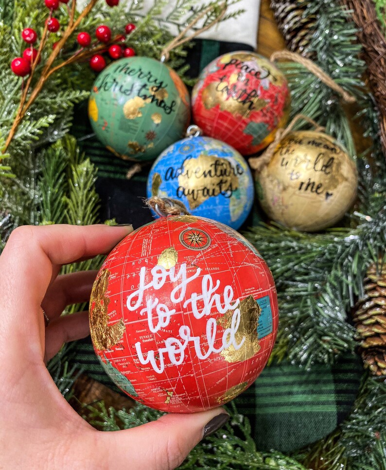 May include: Four red, green, blue, and gold globe ornaments with gold accents and white calligraphy. The ornaments read "Merry Christmas", "Peace on Earth", "Adventure Awaits", and "Joy to the World".