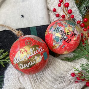 May include: Two red globe ornaments with gold accents. One ornament says "Adventure awaits" and the other says "Mazie Jo 2021".