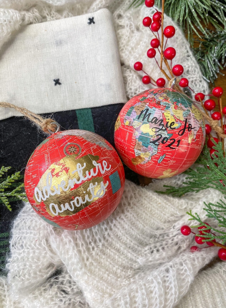 May include: Two red globe ornaments with gold accents. One ornament says "Adventure awaits" and the other says "Mazie Jo 2021".