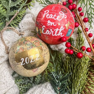 May include: Two globe-shaped Christmas ornaments. One is red with the text "Peace on Earth" and the other is gold with the year "2022" written on it.