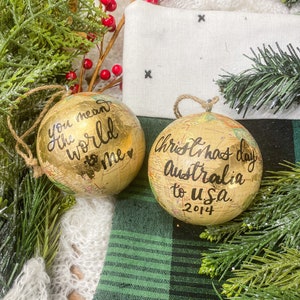 May include: Two gold globe ornaments with black calligraphy. One ornament says "You mean the world to me" with a heart. The other ornament says "Christmas day Australia to U.S.A. 2014".