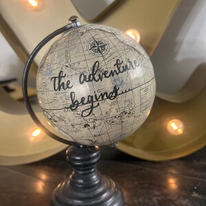 Puede incluir: Un globo terráqueo de estilo vintage con una base negra y un arco de metal negro. El globo presenta un diseño de mapa del mundo con las palabras "the adventure begins" en escritura negra. El fondo incluye una gran letra dorada y luces cálidas.
