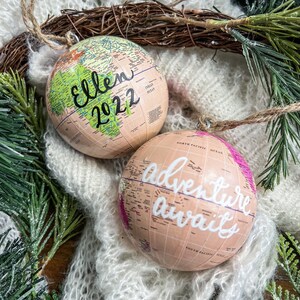 May include: Two globe-shaped Christmas ornaments. One is green and beige with the text "Ellen 2022" written on it. The other is pink and beige with the text "Adventure awaits" written on it.
